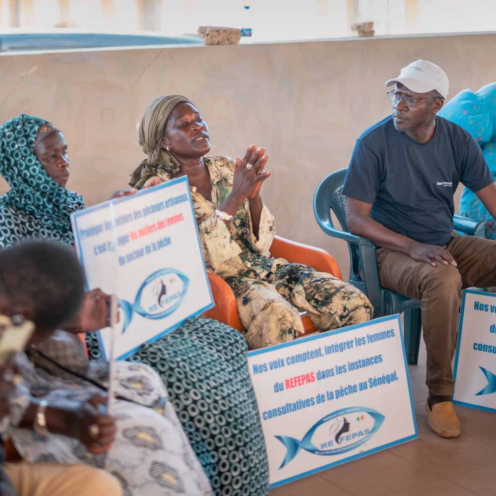 Diaba sitzt auf einem Stuhl und hält ein Schild ihrer Organisation auf einer Versammlung zu Frauenrechte in der Fischerei