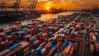 Ein Containerhafen bei Sonnenuntergang mit zahlreichen gestapelten Schiffscontainern, Verladekränen und einem Frachtschiff. Das Bild verdeutlicht die Bedeutung globaler Lieferketten, des internationalen Handels
