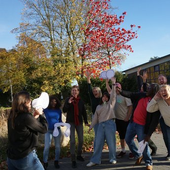 Eine Frau mit einem Plakat, auf dem ein Megafon abgebildet ist, tut so, als rufe sie hinein. Eine Gruppe von Menschen steht im Hintergrund