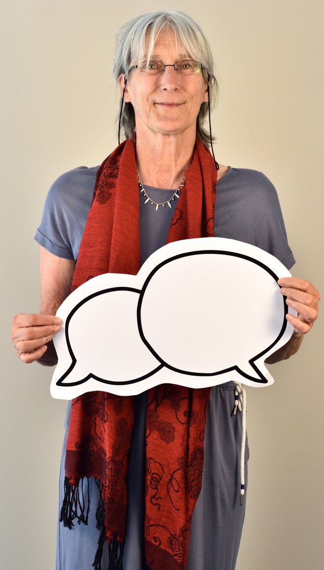 Portrait von Franziska Dickschen, die graue mittellange Haare und eine weiße Haut hat. Die trägt einen roten Schal und ein graues Kleid und hält ein Plakat mit zwei Sprechblasen in den Händen.