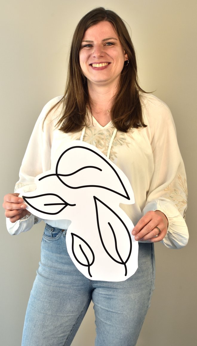 Portrait von Denise Irmscher, die braune lange Haare und weiße Haut hat, eine weiße Bluse trägt und ein Plakat mit gezeichneten Blättern darauf in der Hand hält