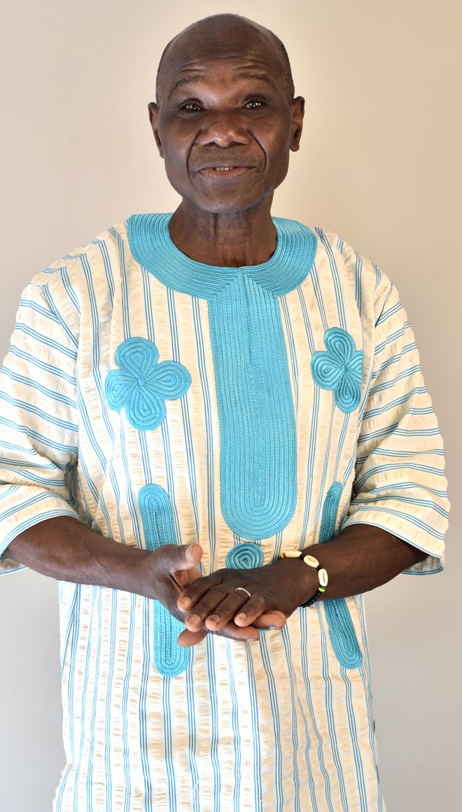 Ein älterer Mensch mit einem blau weißen langen Hemd und schwarzer Hautfarbe schaut in die Kamera und legt die Hände ineinander