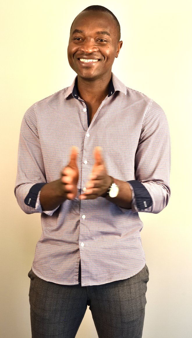 Portrait von Jan Fomboh, der ein rotes Hemd trägt, schwarze Haut hat und ganz kurze Haare. Er lacht und hält die Hände nach vorne