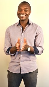 Portrait von Jan Fomboh, der ein rotes Hemd trägt, schwarze Haut hat und ganz kurze Haare. Er lacht und hält die Hände nach vorne