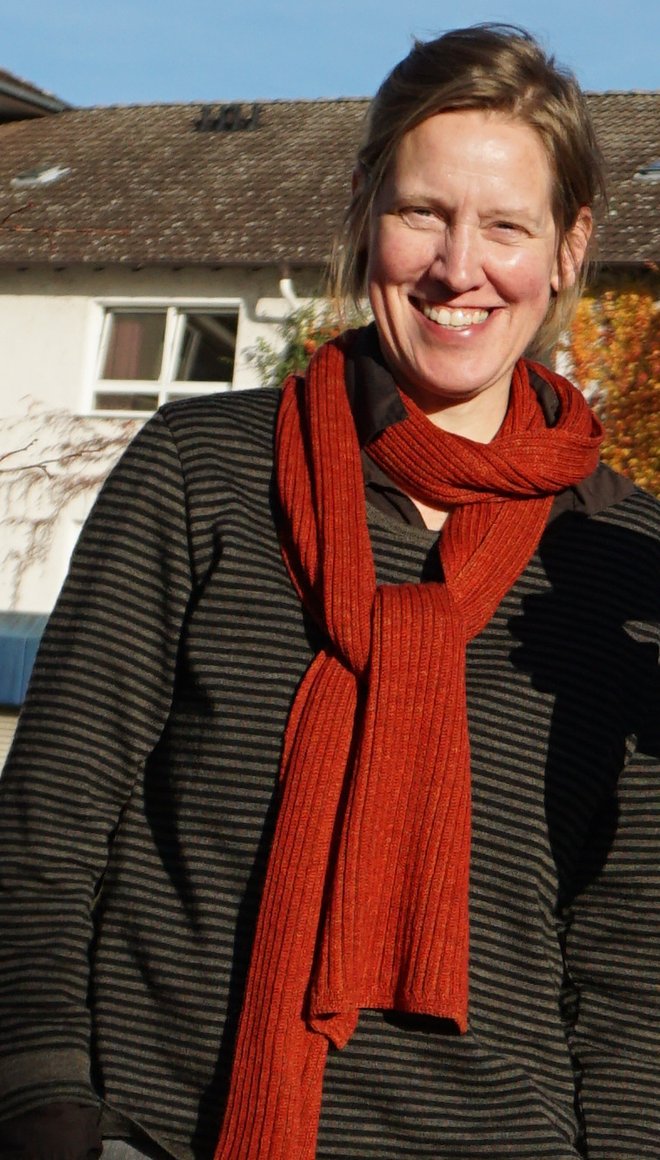Portait von Julia Wältring, die einen blonden Zopf und und weiße Haut hat und einen grauen Pullover und einen roten Schal trägt