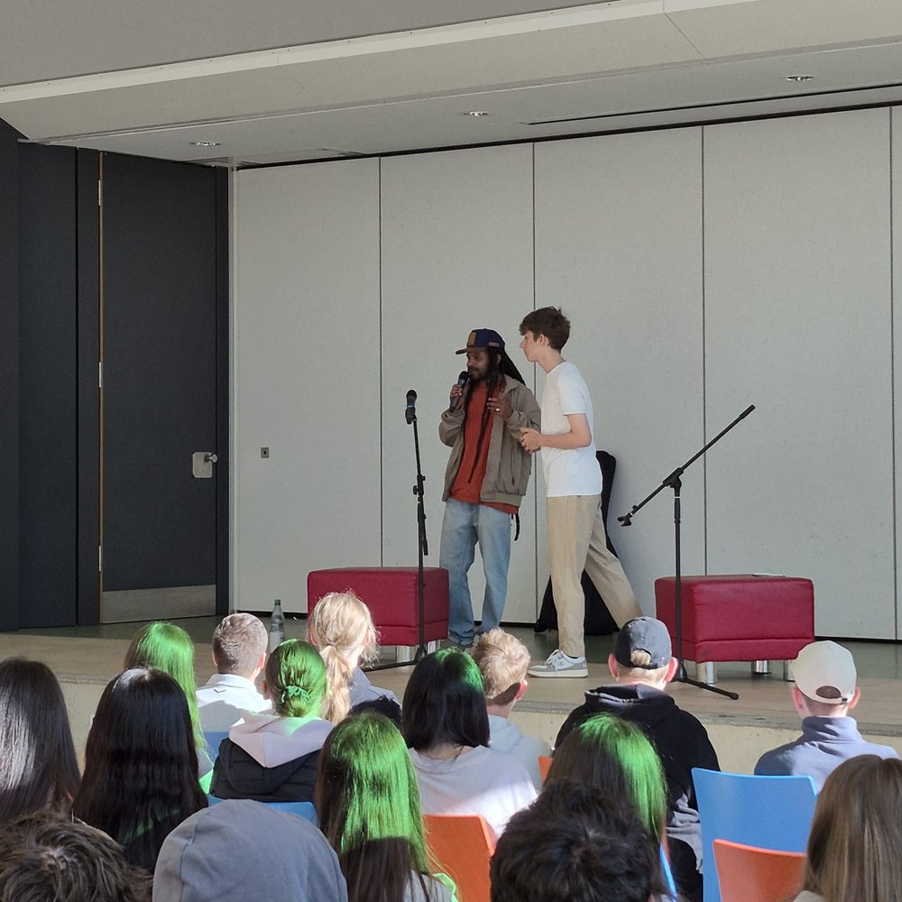 Ein Südgast spricht ins Mikrophone auf der Bühne in einer Hauptschule