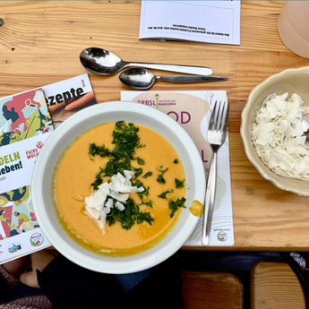 Ein Teller Suppe, ein Programmheft der Fairen Woche in Braunschweig und Besteck liegen auf einem Holztisch.
