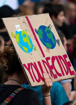 Ein Schild auf einer Demonstration. Auf ihm ist eine Weltkugel nachhaltig und eine zerstört gemalt, darüber der Schriftzug You Decide